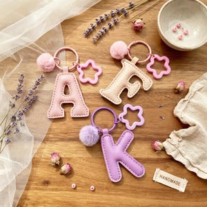 Op de afbeelding: Drie letter-sleutelhangers met de letters A, F en K. Elke sleutelhanger bevat een gewatteerde letterhanger, een bijpassende pluizige pompon en een stervormige bedel bevestigd aan een metalen ring. De A is lichtroze, de F is beige en de K is lavendelkleurig.