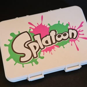 May include: White rectangular case with the word "Splatoon" in a bold, stylized font, set against green and pink paint splatters. The case has a hinged lid and a secure latch, suggesting it's designed for storage.