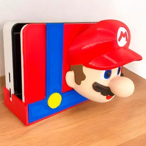 May include: A red, blue, and yellow Mario-themed Nintendo Switch dock. The dock features a 3D-printed Mario head with a red cap and a white "M" on the front. The dock is designed to hold a Nintendo Switch console.