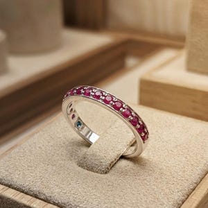 May include: A silver-toned band ring featuring a continuous row of small, round, vibrant pinkish-red gemstones set along the top half. The metal band has a polished finish, and a single small blue stone is visible on the inner side of the ring.