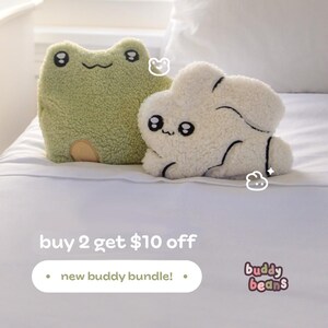 May include: Two plush stuffed animal pillows, one shaped like a green frog and the other like a white cloud. Both have embroidered black eyes and smiles. The frog has a tan belly. The text "buy 2 get $10 off" and "new buddy bundle!" is also visible.