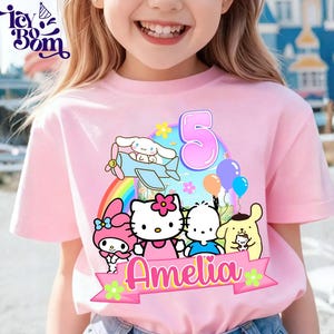 May include: Pink short-sleeved t-shirt featuring a colorful graphic of cartoon characters, a rainbow, and the number 5. The name "Amelia" is displayed on a banner. The shirt is designed for a child.