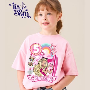 May include: A pink t-shirt featuring a Barbie graphic, a number 5, and the name "Madelyn". The design includes a rainbow, stars, and surfboards. The shirt is worn by a young girl.