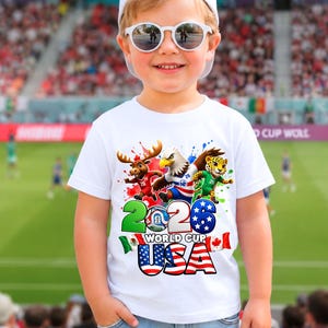 May include: A young child wearing a white t-shirt with a colorful graphic design. The shirt features the text "2026 World Cup USA" along with cartoon animal characters and the American flag. The child is also wearing sunglasses and a white cap.