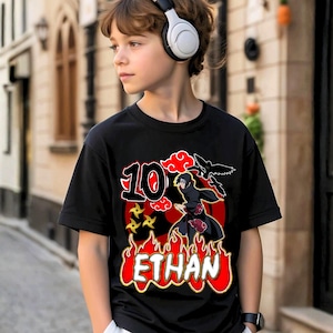 May include: Black t-shirt with a red and yellow graphic design featuring the number 10, a character illustration, and the name "ETHAN" in flames. The shirt is worn by a person wearing white headphones.