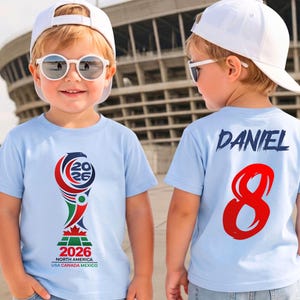 May include: A light blue t-shirt featuring a 2026 North America soccer graphic on the front and the name DANIEL with the number 8 in red on the back. The graphic includes a stylized trophy design with the text 2026 NORTH AMERICA USA CANADA MEXICO.