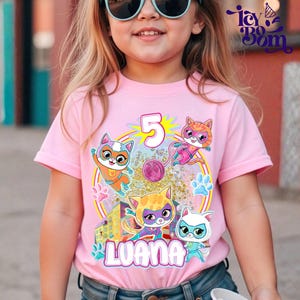 May include: A pink t-shirt with a cartoon cat design, the number 5, and the name "Luana". The graphic includes superhero cats, paw prints, and a ball of yarn. The t-shirt is worn by a young girl, and is a children's garment.