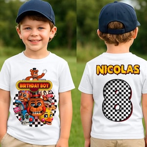 May include: White t-shirt with a colourful graphic featuring cartoon characters and the text "BIRTHDAY BOY." The back of the shirt displays the name "NICOLAS" and a checkered number 8. The child is wearing a blue baseball cap.