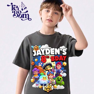 May include: A dark gray t-shirt featuring a colorful graphic with the text "JAYDEN'S 8TH BDAY" and cartoon characters. The shirt is worn by a person wearing a baseball cap.