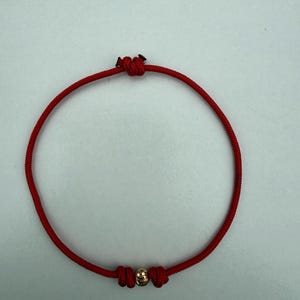 May include: Red string bracelet with a gold-colored bead. The bracelet is knotted at the ends and around the bead. The bracelet is a simple, minimalist design.