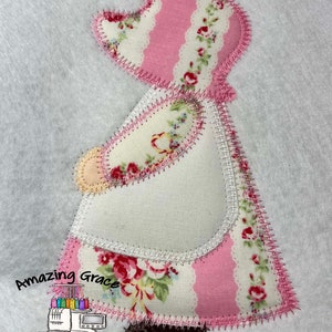 May include: A pink and white fabric applique of a girl with a floral print dress and a brown shoe. The applique is stitched with a white and pink thread. The text "Amazing Grace Embroidery Designs" is below the applique.