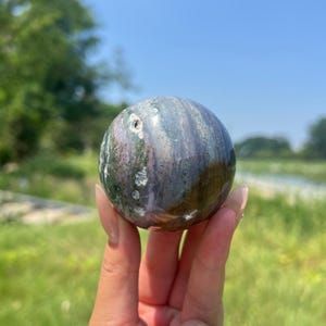 Puede incluir: Una piedra esférica pulida que presenta un patrón de remolinos en tonos violeta apagado, verde oscuro y gris con pequeñas inclusiones blancas. La superficie del orbe es lisa y reflectante, mostrando variaciones minerales naturales.