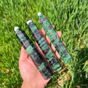 Può includere: Tre bacchette di cristallo di rubino zoisite lucide. Le bacchette sono verdi con venature nere e rosa. Ogni bacchetta ha una punta bianca. Le bacchette sono tenute in mano su uno sfondo di erba verde.
