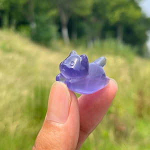 Puede incluir: Una pequeña figura de piedra preciosa tallada, translúcida y de color violeta, con forma de gato. El gato aparece en posición de descanso con las orejas hacia arriba y la cola curvada hacia atrás, con una superficie lisa y pulida.