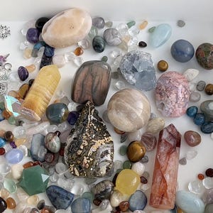 May include: A collection of various polished gemstones and crystals in a white tray. The assortment includes quartz, amethyst, and other colorful stones in shades of pink, blue, yellow, and brown. Some are tumbled, others are faceted.