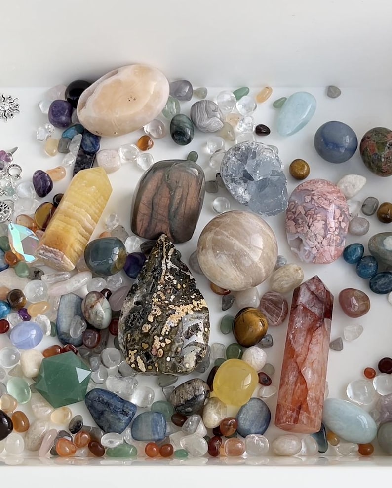 May include: A collection of various polished gemstones and crystals in a white tray. The assortment includes quartz, amethyst, and other colorful stones in shades of pink, blue, yellow, and brown. Some are tumbled, others are faceted.