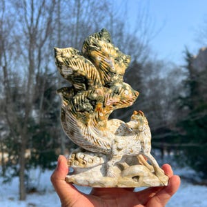 Può includere: Scultura in pietra scolpita raffigurante un lupo e una creatura multicefala. La scultura è principalmente beige, con accenti verdi e marroni. Il lupo è raffigurato in piedi su una base, mentre l'altra creatura ha più teste.