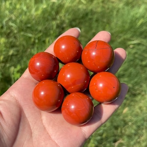 May include: Seven polished red jasper spheres arranged in a cluster. Each stone is spherical, smooth, and features a deep, solid red colour with subtle natural variations, including faint dark veining and small black speckles on the surface.