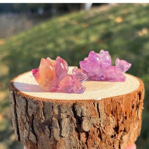 May include: Two small, carved fox figurines made of translucent, polished purple and orange fluorite crystal. The foxes are depicted in a curled, resting position with detailed tails, showcasing natural colour variations and smooth, reflective surfaces.