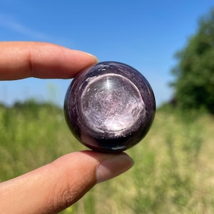 Peut inclure: Une pierre précieuse sphérique polie d'une teinte violet foncé et pourpre profond. La surface de la pierre présente un éclat métallique réfléchissant avec des marques de polissage circulaires visibles et des stries internes captant la lumière.
