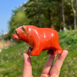 Op de afbeelding: Een gepolijst rood jaspis stenen beeldje in de vorm van een beer. Het figuurtje heeft een glad, glanzend oppervlak met natuurlijke minerale variaties, waaronder donkerdere roodbruine strepen en een solide, afgeronde vorm.
