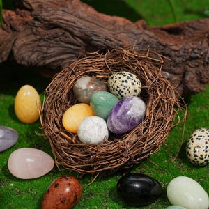 May include: A collection of colorful gemstone eggs in a woven nest, set against a backdrop of green grass and a piece of driftwood. The eggs vary in color, including yellow, purple, green, and speckled patterns, creating a natural and decorative display.