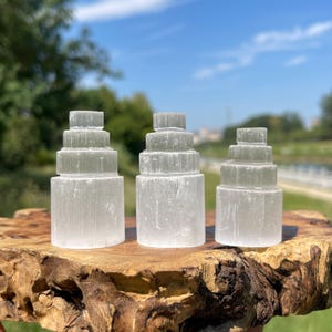 Può includere: Tre torri di selenite bianca, ciascuna intagliata in una forma cilindrica a gradini con quattro livelli. Le strutture minerali fibrose e traslucide sono disposte fianco a fianco, mostrando una finitura liscia e un aspetto cristallino luminoso.