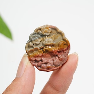 Può includere: Un ciondolo circolare in pietra intagliata raffigurante una volpe in rilievo circondata da motivi floreali. La pietra presenta una tavolozza di colori naturali composta da marroni terrosi, rossi profondi e grigi tenui con finitura lucida.