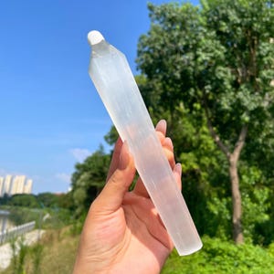 May include: A long, translucent white selenite crystal wand with a faceted, pointed tip. The crystal has a smooth, fibrous texture and a cylindrical shape, catching the sunlight to display a soft, glowing appearance throughout its length.