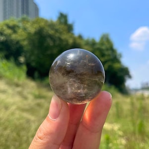 Puede incluir: Una esfera de cristal de cuarzo ahumado pulido con un tono marrón oscuro translúcido. La piedra presenta inclusiones naturales y patrones nublados en toda su superficie redondeada, reflejando la luz con un acabado suave y brillante.