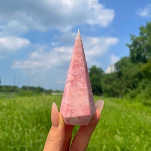 May include: A polished pink crystal tower with a pointed top and a hexagonal base. The stone features a marbled texture with varying shades of light and soft pink throughout its surface, displaying natural veining and a smooth, reflective finish.