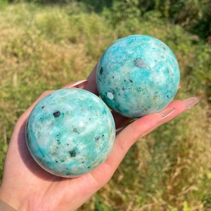 May include: Two polished spherical stones featuring a vibrant turquoise and teal colour palette with mottled white veining and small dark mineral inclusions throughout the smooth, rounded surfaces of these natural gemstone spheres.