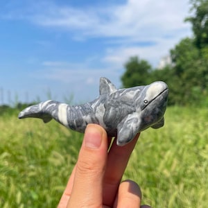 Puede incluir: Una figura de piedra tallada a mano con forma de orca, que presenta una superficie pulida con un patrón jaspeado de remolinos de color gris oscuro, gris claro y blanco, una aleta dorsal prominente y un cuerpo liso y redondeado con cola.
