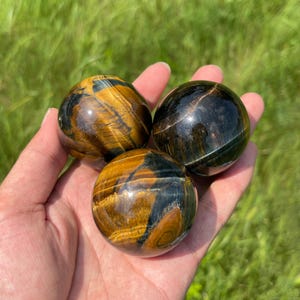 May include: Three polished spherical stones featuring a chatoyant tiger's eye pattern with alternating bands of golden-brown and deep black. The smooth, rounded surfaces exhibit a reflective lustre, showcasing the natural mineral striations.