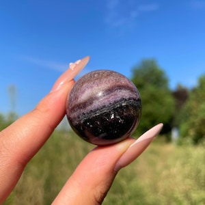 May include: A polished spherical stone featuring a gradient of colours ranging from deep black at the base to layers of dark purple, lavender, and white bands across the top. The surface is smooth and reflective, showing natural mineral inclusions.