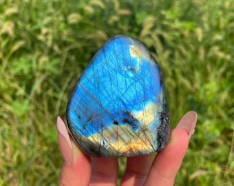 May include: A polished labradorite stone featuring a vibrant blue flash across its surface, with visible internal fractures and golden-yellow undertones. The stone has a smooth, rounded triangular shape with a dark, natural grey edge.