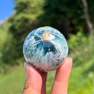 Könnte beinhalten: Eine klare, kugelförmige Kristallkugel mit einem blau-weißen, federartigen Muster im Inneren. Die Kugel wird in einer Hand vor einem verschwommenen grünen Hintergrund gehalten. Die Kristallkugel hat einen kleinen, hellbraunen Einschluss oben.