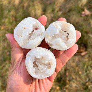 May include: Three white geode stones with crystal interiors are held in a hand. The geodes have a rough, textured exterior and a hollowed-out centre filled with sparkling crystals. The stones are round and vary slightly in size.