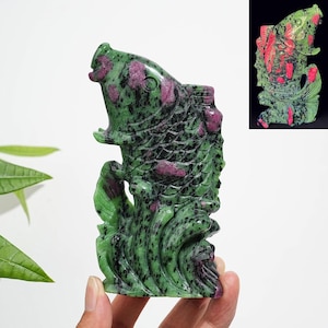May include: A hand-carved zoisite sculpture featuring a fish leaping from stylized waves. The stone displays a mottled green base with prominent inclusions of deep magenta ruby throughout the body and fins, creating a textured, natural appearance.