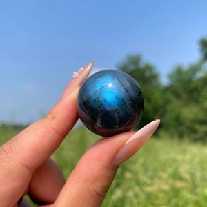 May include: A polished labradorite sphere displaying a deep dark grey base colour with a vibrant, iridescent blue flash across its surface. The stone is spherical in shape, featuring natural mineral inclusions and a smooth, reflective finish.