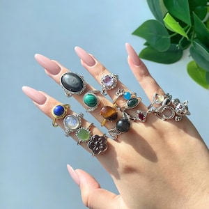 Può includere: Una collezione di anelli color argento con varie pietre preziose tra cui lapislazzuli, pietra di luna, malachite, occhio di tigre, ametista e onice nero. Gli anelli presentano forme diverse come ovali, cerchi e un design a forma di gatto.