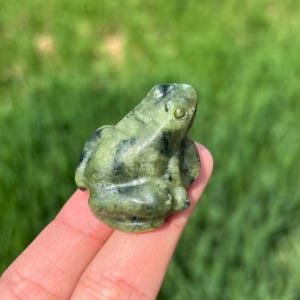 Op de afbeelding: Een kleine, groene kikkerfiguur met zwarte markeringen. De kikker is uit steen gesneden en heeft een glad, gepolijst oppervlak. Het beeldje wordt in de hand van een persoon gehouden tegen een vage groene achtergrond.