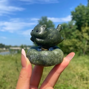 May include: A dark green stone carving of a small, rounded creature with two black eyes, positioned on top of a smooth, circular stone base. The figure has a stout body, small ears, and a short tail, with a polished texture throughout the piece.