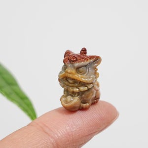 May include: A small, intricately carved stone figurine of a mythical lion creature. The carving features a reddish-brown top section transitioning into a tan and brown base, with detailed facial features, pointed ears, and a small knob on the head.