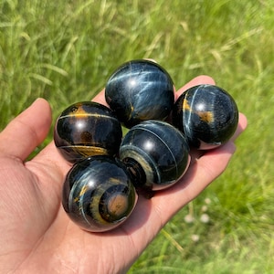 May include: Five polished spheres made of blue tiger's eye gemstone. Each sphere features a smooth, reflective surface with distinct chatoyant bands of deep navy blue, charcoal grey, and golden-brown mineral inclusions that create a shimmering effect.