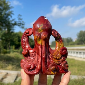 Könnte beinhalten: Eine geschnitzte Oktopus-Figur aus Stein mit einer glatten, polierten Oberfläche und tiefroten, orangefarbenen und gelben Marmormustern. Die Skulptur zeigt detaillierte Tentakel mit kreisförmigen Saugnapfstrukturen und einen abgerundeten Kopf.