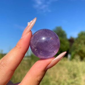 Peut inclure: Une sphère lisse et polie en cristal d'améthyste violet translucide. La pierre présente des inclusions naturelles et des variations d'intensité de couleur, reflétant la lumière sur sa surface arrondie tout en affichant des teintes violettes.