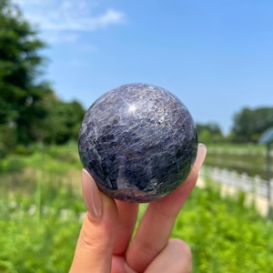 May include: A polished spherical stone with a deep purple and dark blue colour palette. The surface of the sphere displays natural crystalline textures, internal fractures, and subtle reflective highlights across its rounded, smooth, and solid form.
