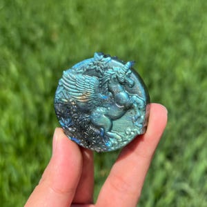 May include: A carved, round, iridescent stone featuring a winged unicorn design. The stone has shades of blue, green, and gold. The sculpture is held in a hand against a blurred green background.
