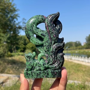 May include: A carved zoisite sculpture featuring a multi-tailed fox figure with prominent green and black speckled patterns and streaks of deep magenta. The polished stone ornament displays intricate curves and a smooth, reflective surface texture.